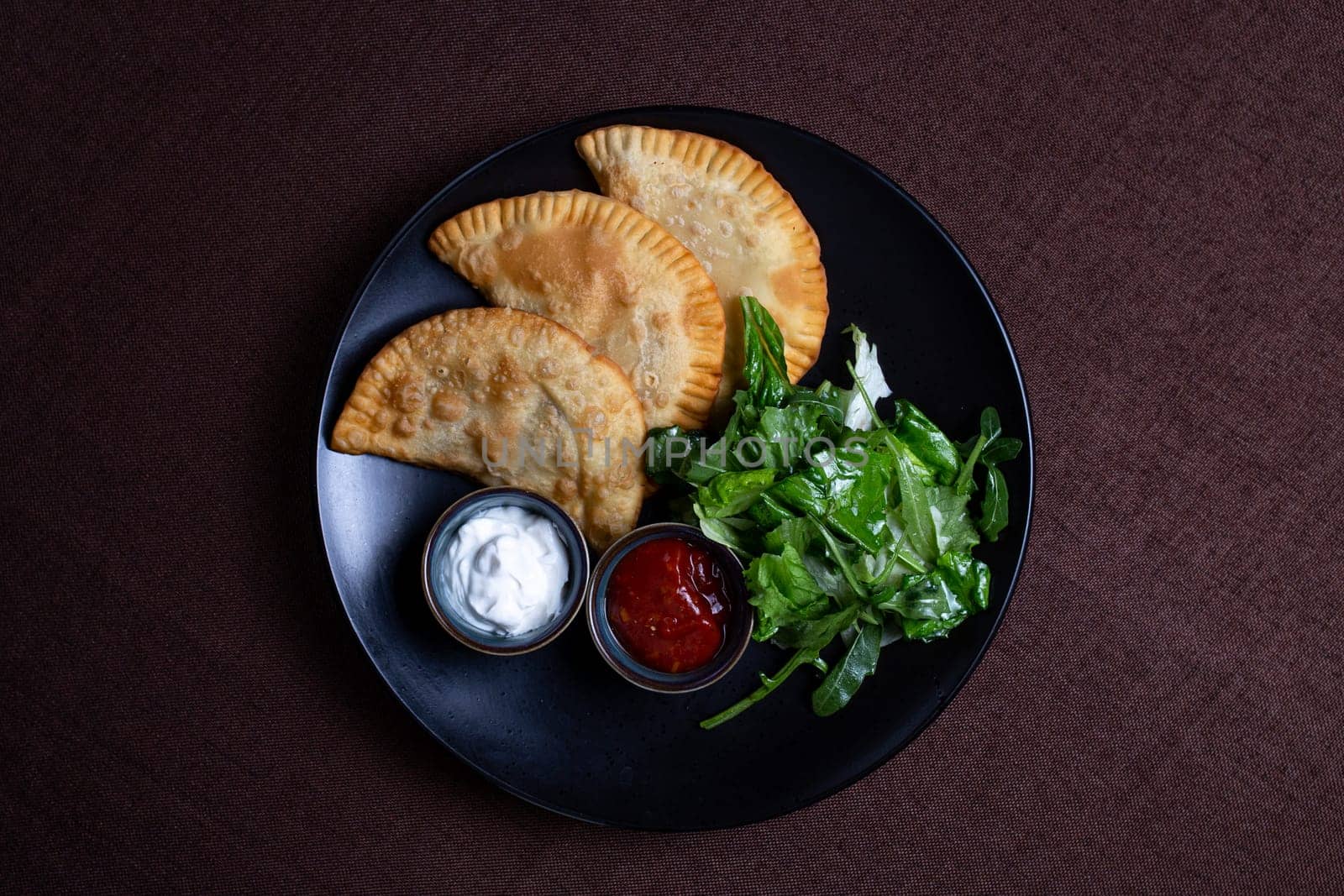 top view fried chebureks with sauce and herbs on a black plate.