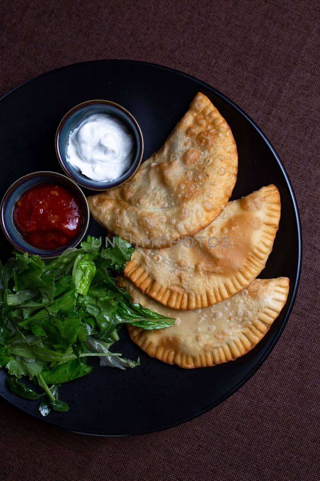 top view fried chebureks with sauce and herbs on a black plate by Pukhovskiy