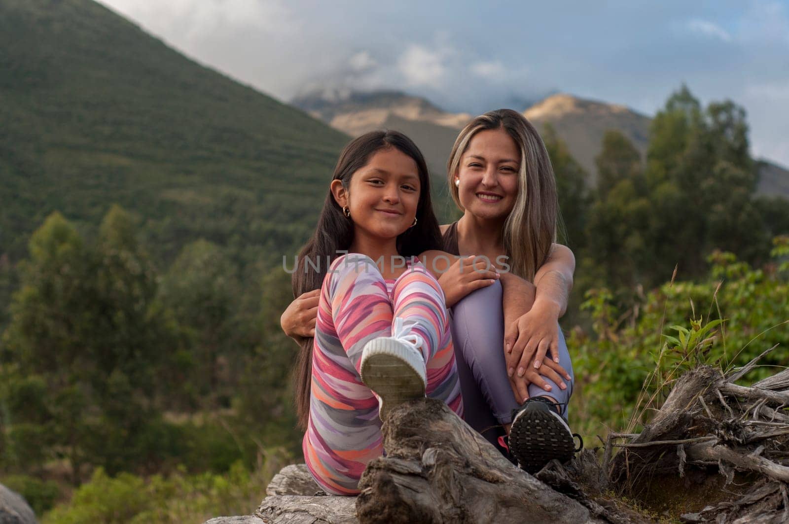 mother and daughter sitting on the trunk of a tree looking at the camera in the middle of nature by Raulmartin