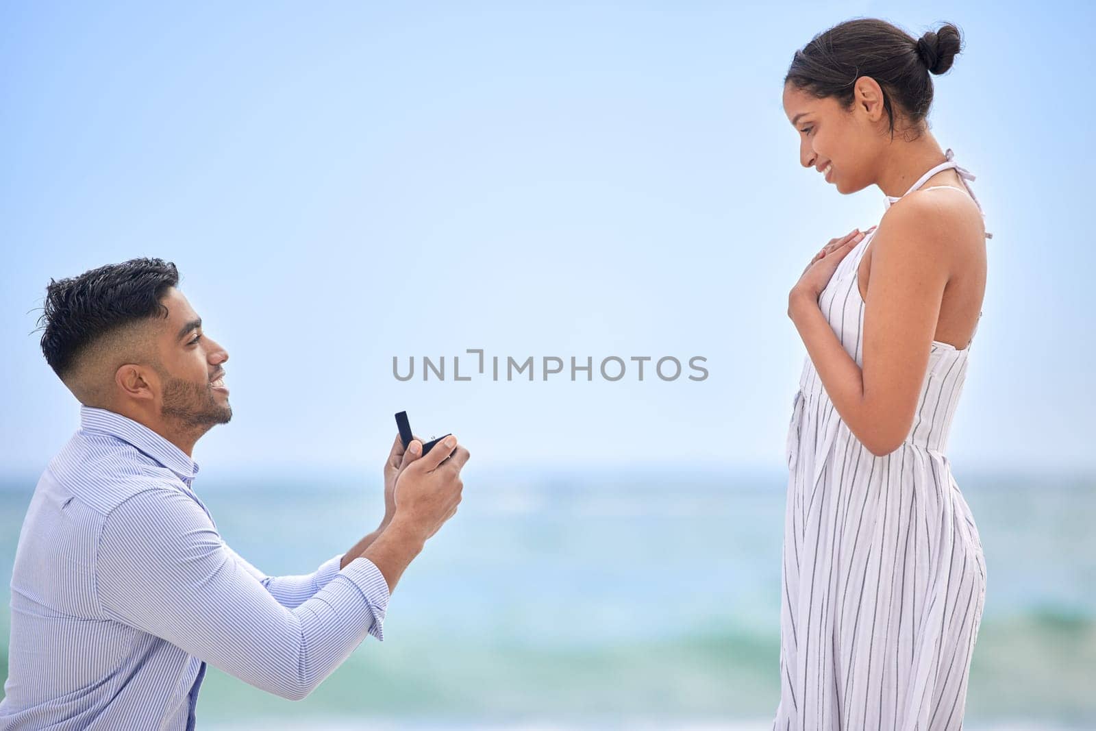 Engagement, proposal and romance with a couple on the beach for a milestone on a blue sky background. Love, summer or nature with a man down on one knee to ask his girlfriend the marriage question by YuriArcurs