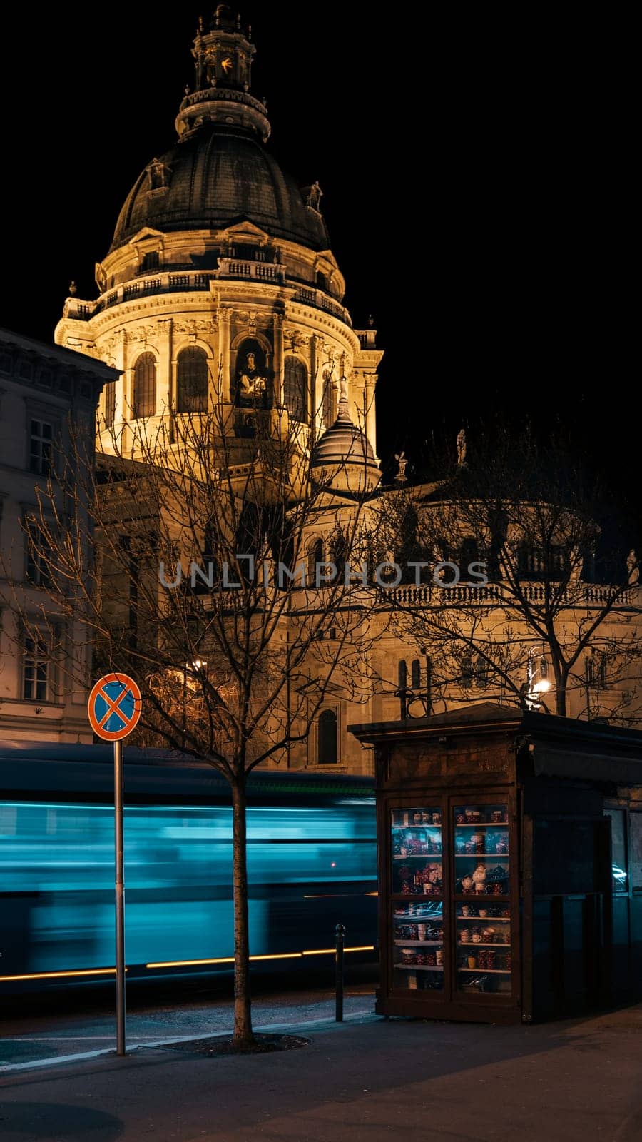 An illuminated Hungarian city comes alive in the evening. Landmarks including a cathedral and newsstand are visible alongside blurry buses and traffic.