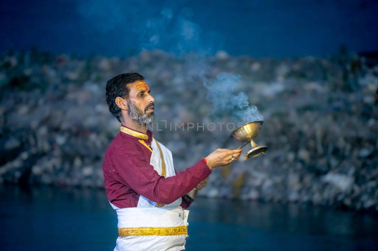 Rishikesh, India - 29th Apr'23: : Priest in red and white praying to the holy river ganga with a lit oil diya lamp giving off smoke as part of the evening aarti prayer performance at triveni ghat