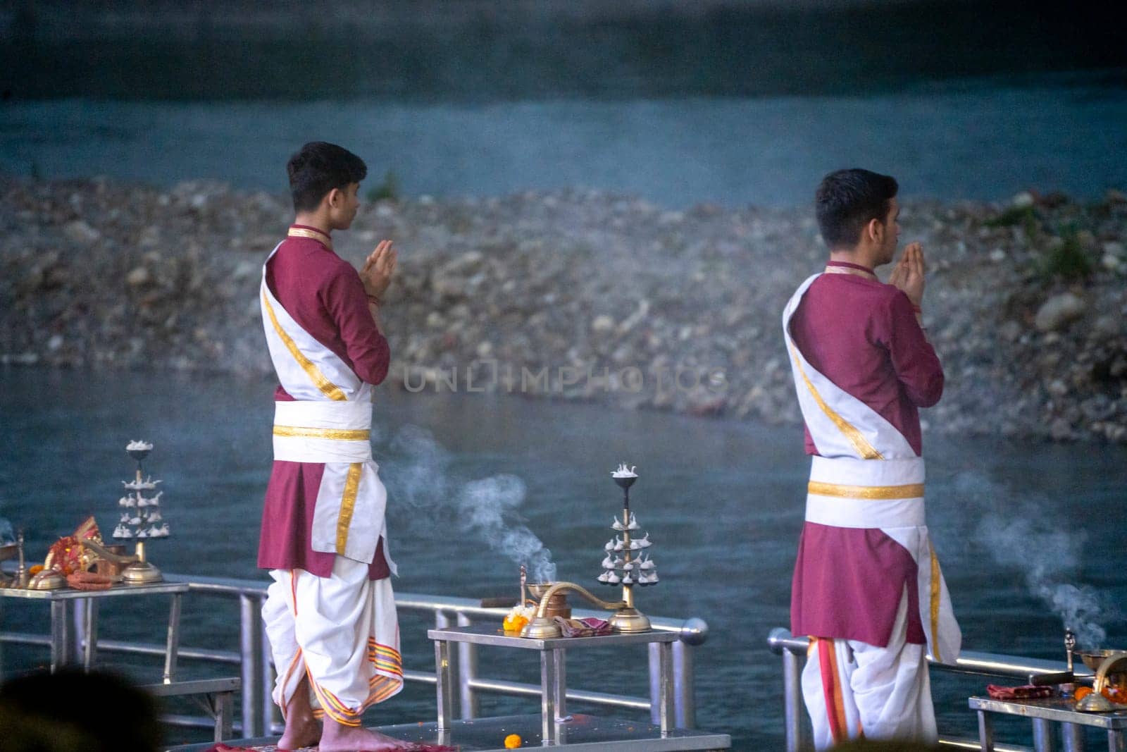 Priest in red and white chanting hymns to the holy river ganga with a lit oil diya lamp giving off smoke as part of the evening aarti prayer performance at triveni ghat
