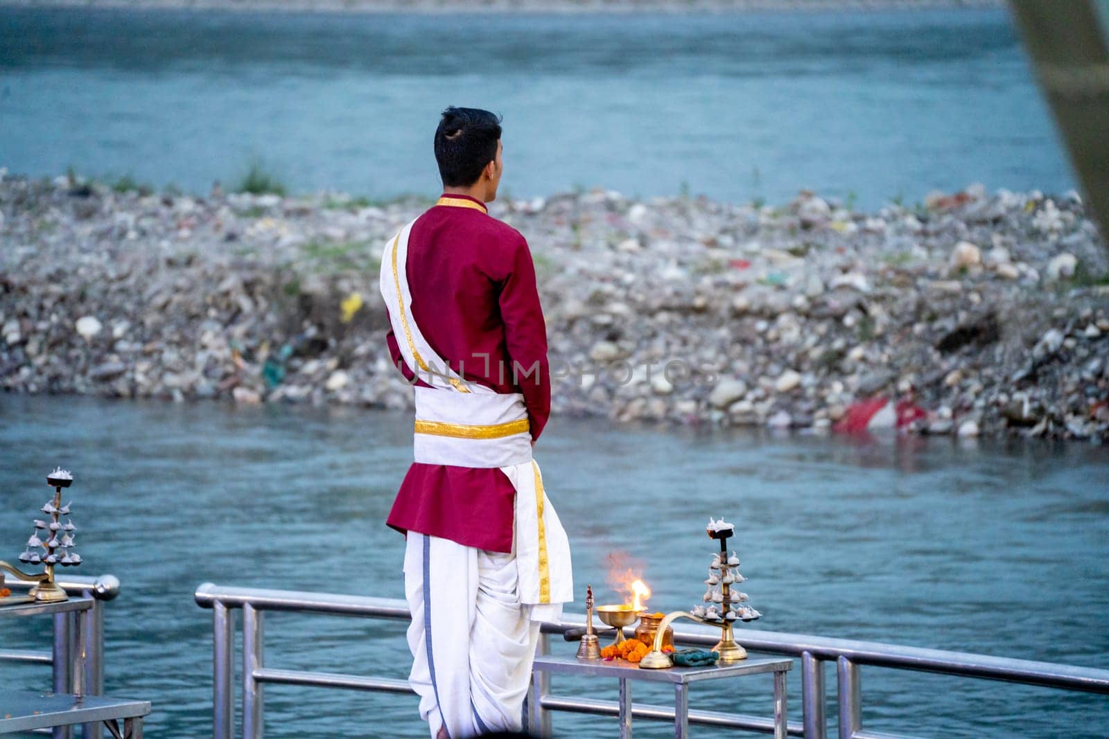 Priest in red and white chanting hymns to the holy river ganga with a lit oil diya lamp giving off smoke as part of the evening aarti prayer performance at triveni ghat