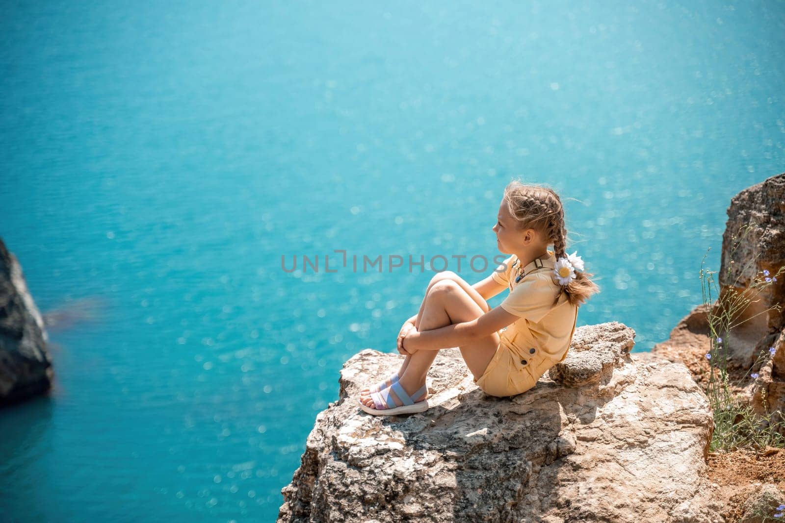 Happy girl perched atop a high rock above the sea, wearing a yellow jumpsuit and braided hair, signifying the concept of summer vacation at the beach
