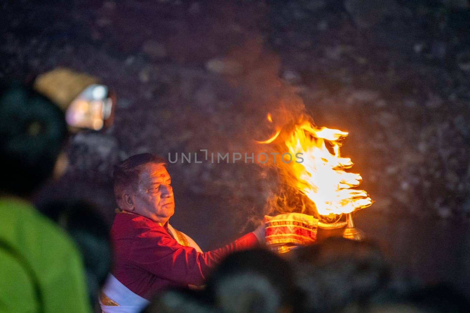 Rishikesh, India - 29th Apr'23: Priest in red and white praying to the holy river ganga with a lit oil diya lamp giving off smoke as part of the evening aarti prayer performance at triveni ghat India