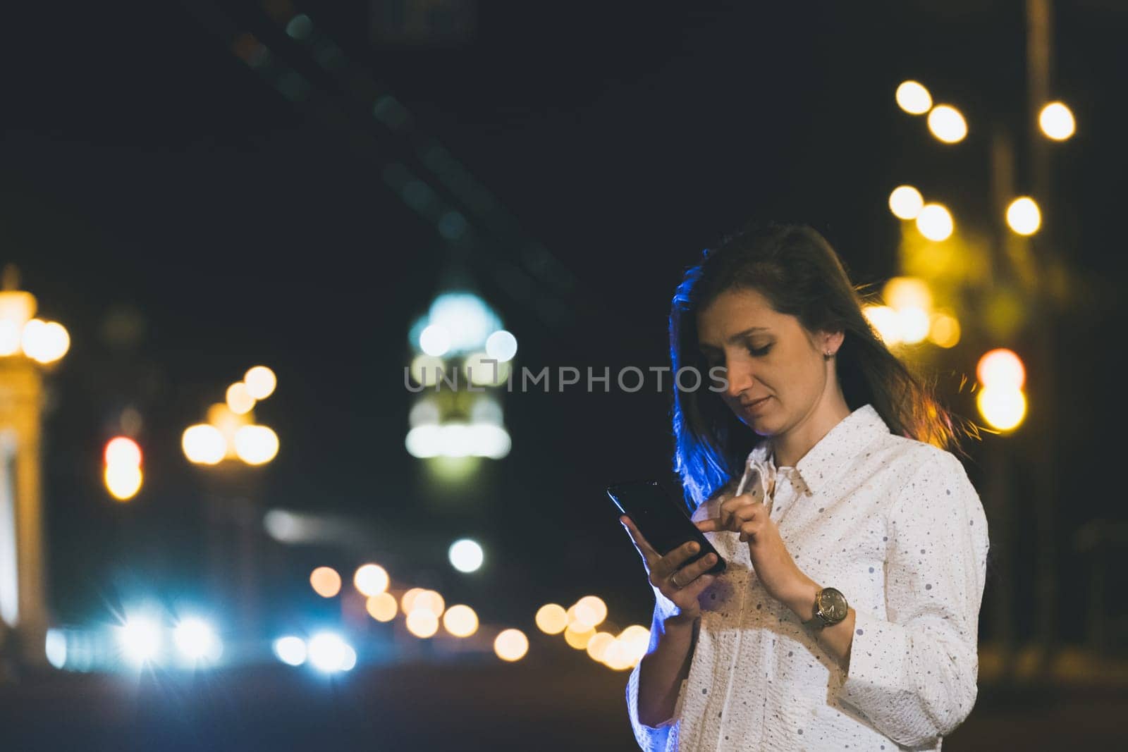 attractive girl in white suit using smartphone in night, light reflection with copy space area for advertising, young woman smiling with mobile in hands