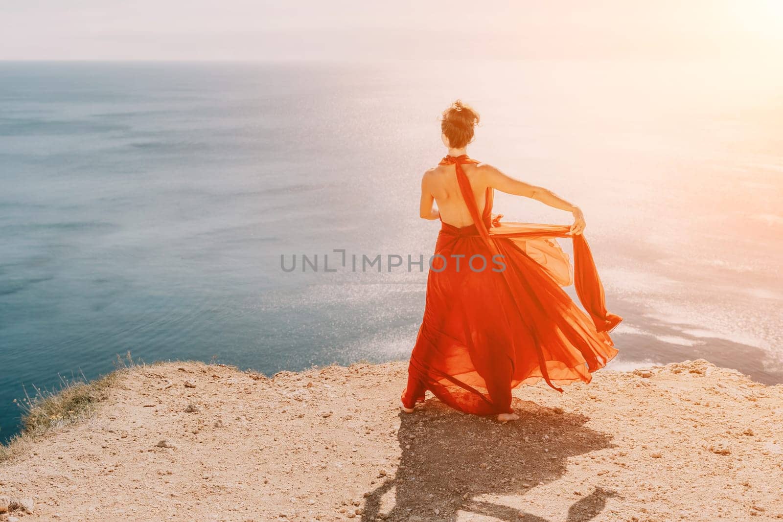 Side view a Young beautiful sensual woman in a red long dress posing on a rock high above the sea during sunrise. Girl on the nature on blue sky background. Fashion photo.