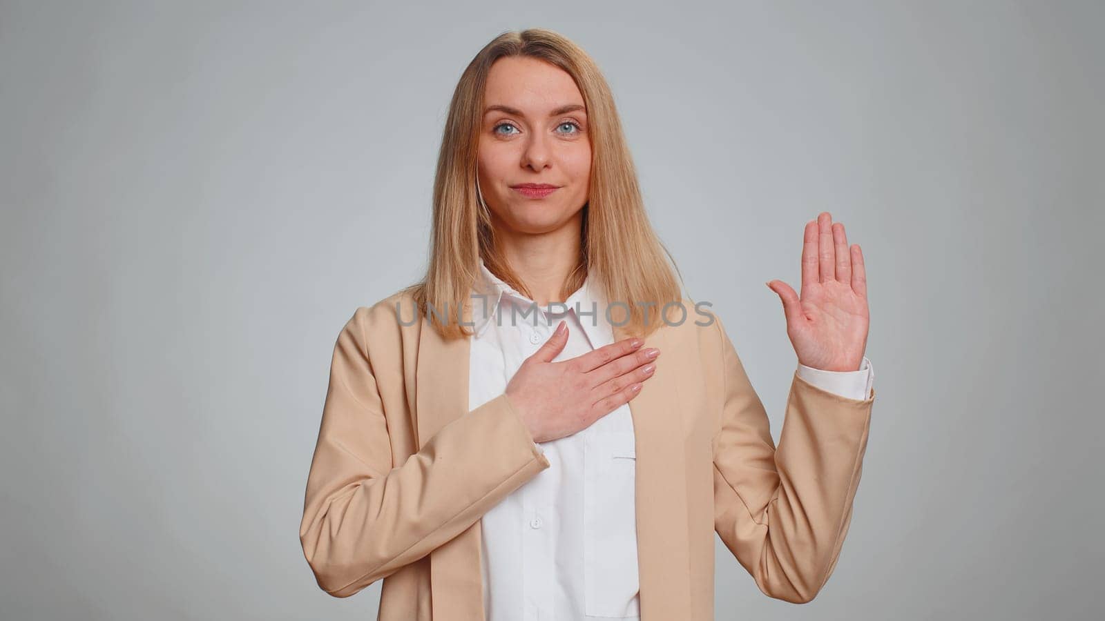 I swear to be honest. Sincere responsible business woman raising hand to take oath, promising to be honest and to tell truth, keeping hand on chest. Secretary girl isolated on gray studio background