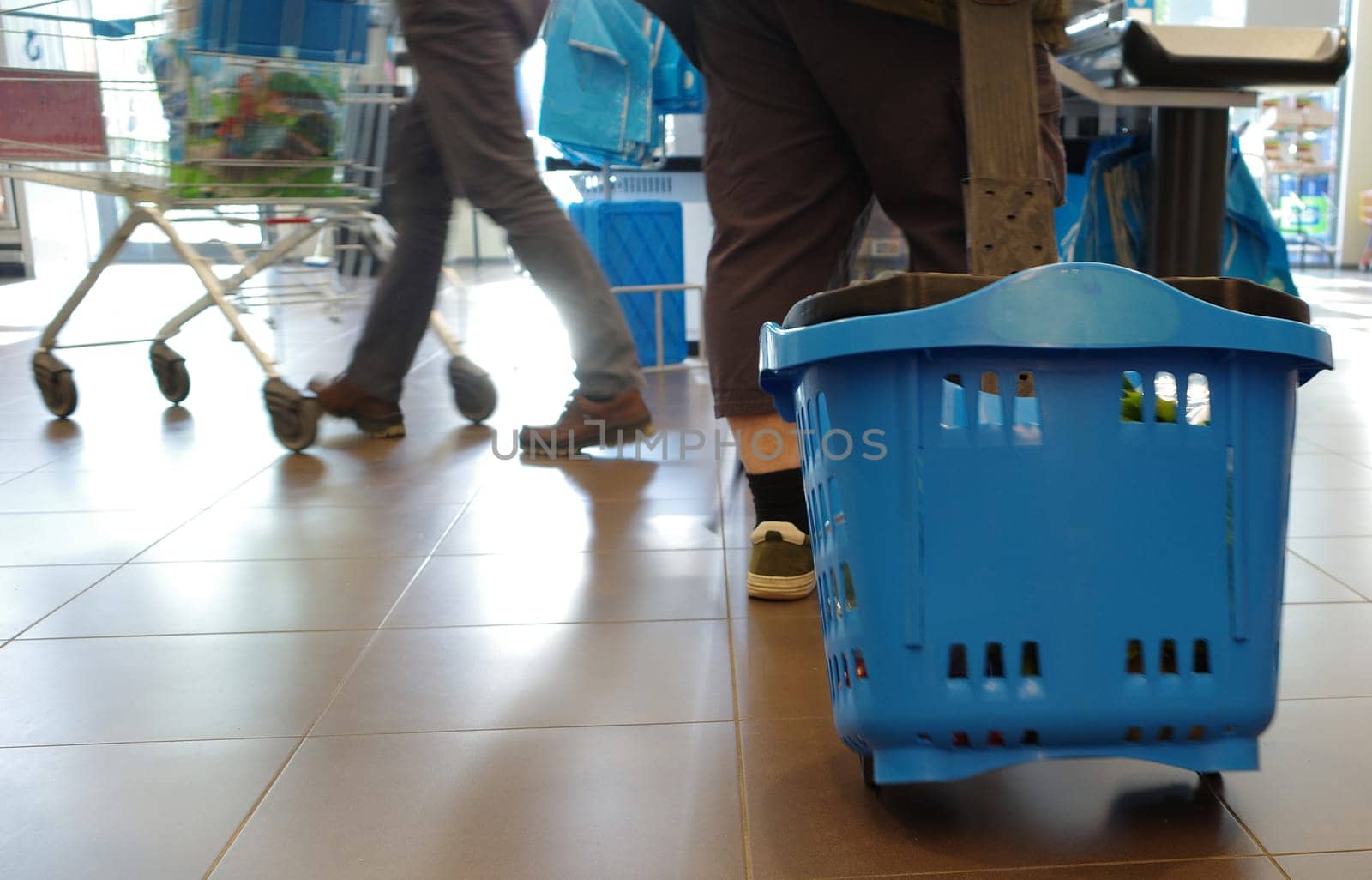 The legs of a man who is shopping. He pulls a blue plastic shopping basket on rollers behind him. In the background are the legs of a person with a shopping cart visible