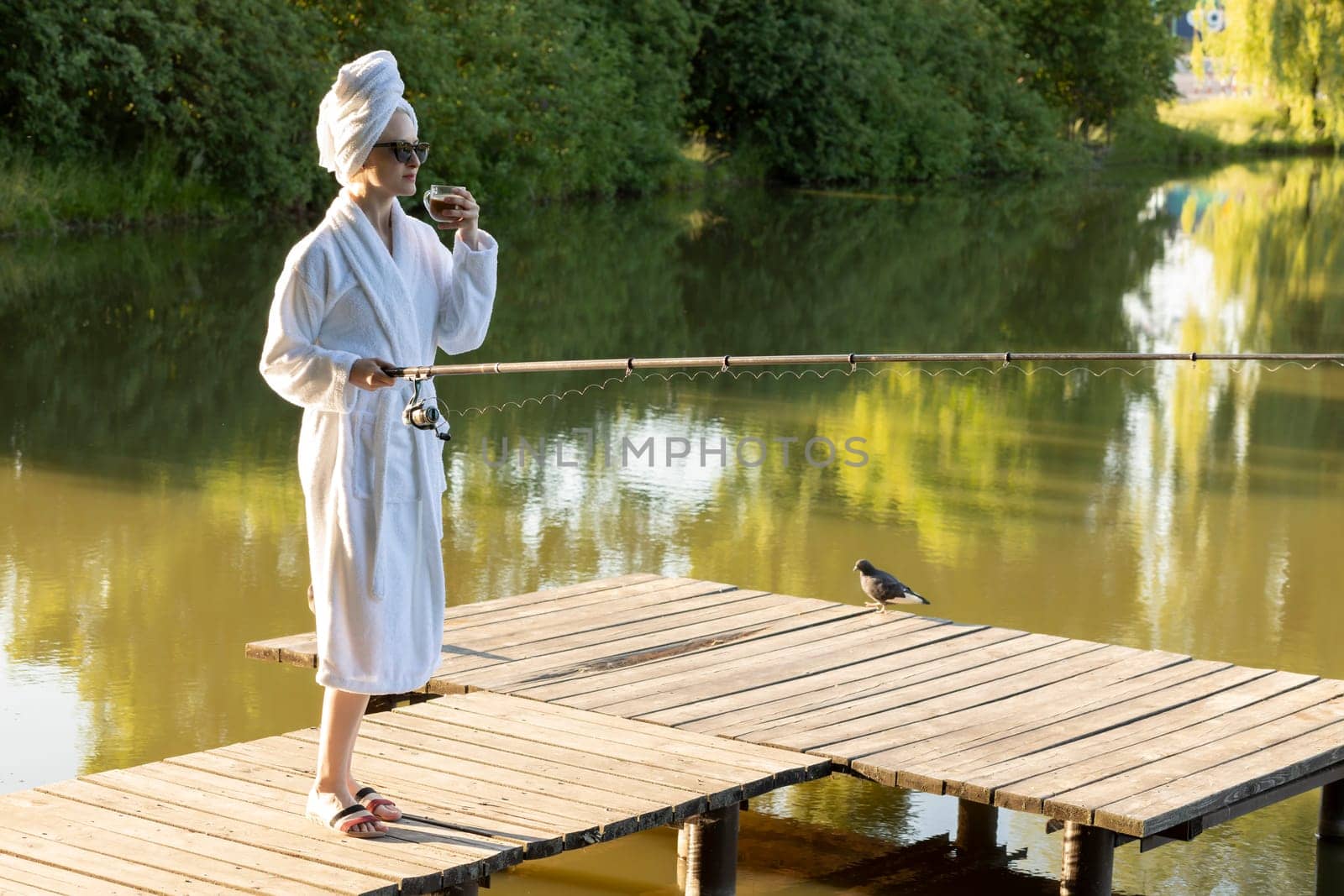 Caucasian Woman In White Robe And Towel Is Fishing Holding Spinning Rod On Pier Of Lake Early In The Morning, Green Trees, Nature On Background. Hobby, Leisure Activity, Relaxation. Horizontal