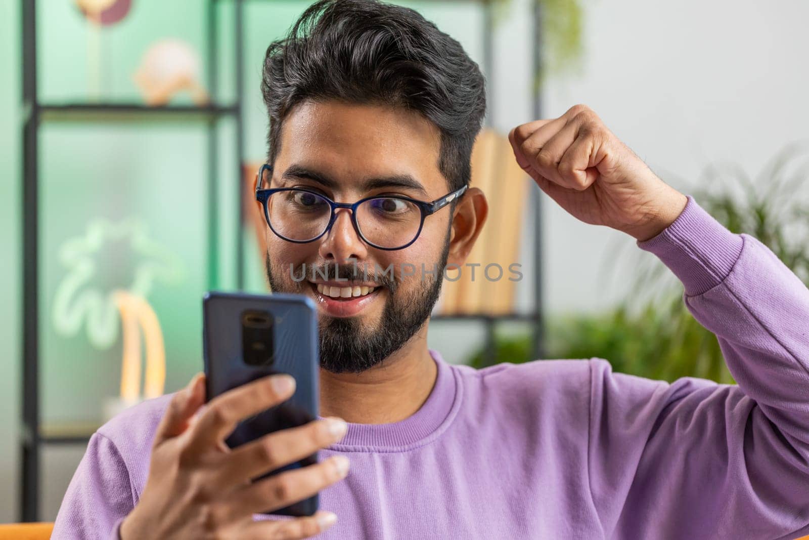 Happy indian man use mobile smartphone typing browsing say Wow yes found out great big win good news celebrate lottery jackpot doing winner gesture. Hindu young guy at home in room sitting on sofa