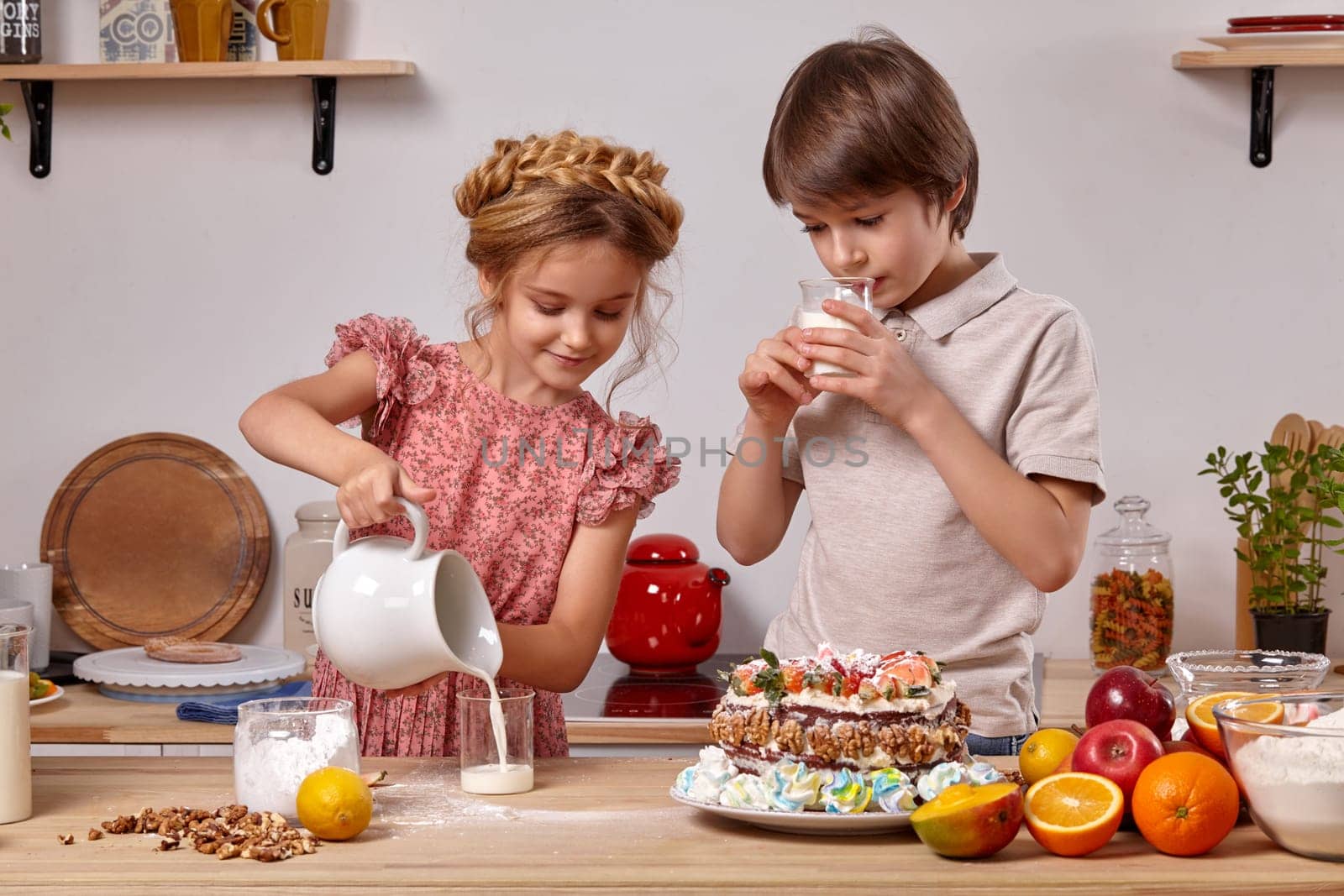 Brunette boy dressed in a light t-shirt and jeans and a beautiful girl wearing in a pink dress are making a cake at a kitchen, against a white wall with shelves on it. Girl is pouring some milk in a glass and boy is drinking his milk.