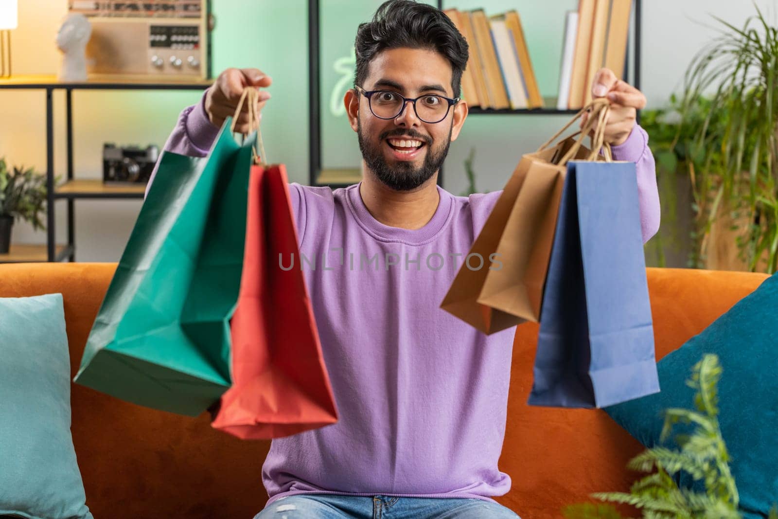 Young indian man happy shopaholic consumer came back home after shopping sale with bags. Hindu guy satisfied received parcels purchase from online order at home apartment room on sofa. Vertical