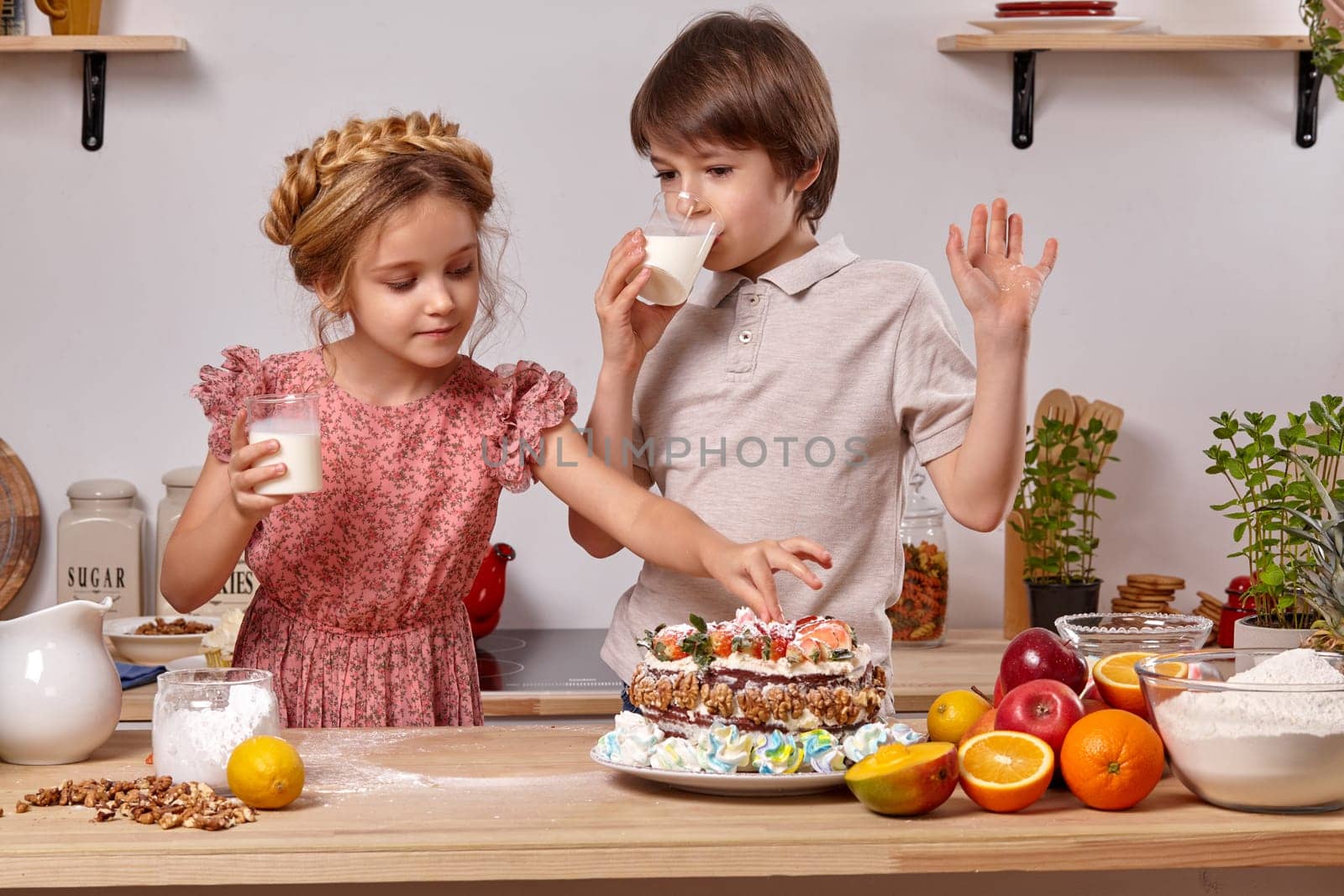 Kid dressed in a light t-shirt and jeans and a beautiful little lady wearing in a pink dress are making a cake at a kitchen, against a white wall with shelves on it. Boy is drinking milk and girl is touching the cake.