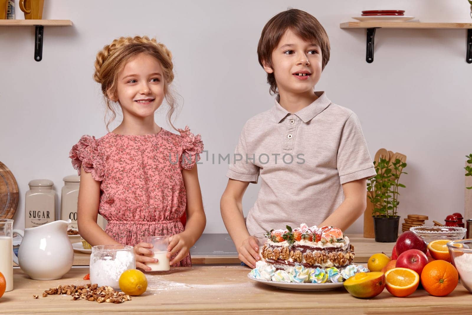Little handsome boy dressed in a light t-shirt and jeans and a charming girl wearing in a pink dress are making a cake at a kitchen, against a white wall with shelves on it. They are smiling and looking away.