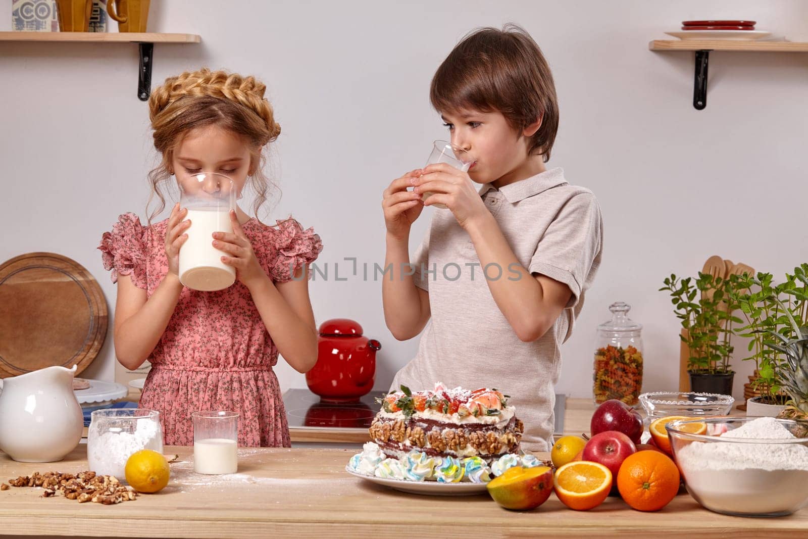 Pretty boy dressed in a light t-shirt and jeans and a funny girl wearing in a pink dress are making a cake at a kitchen, against a white wall with shelves on it. Girl is drinking milk from a big jug and boy is looking at her.