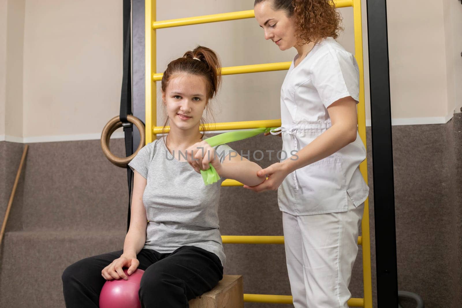 Physician Helps Disabled Child Do Physical Exercises In Rehabilitation Room. Kid With Special Needs. Rehabilitation. Smiling Girl Pulls Fitness Rubber Bands. Horizontal plane. High quality photo