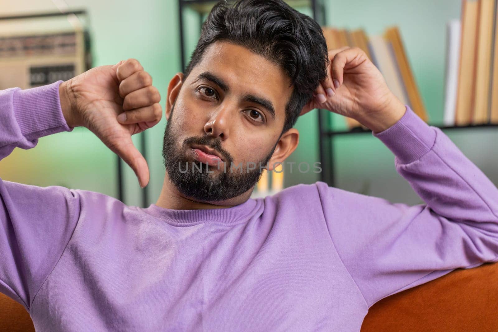 Dislike. Upset indian man showing thumbs down sign gesture, expressing discontent, disapproval dissatisfied bad work at modern home apartment indoors. Displeased hindu guy in living room on sofa