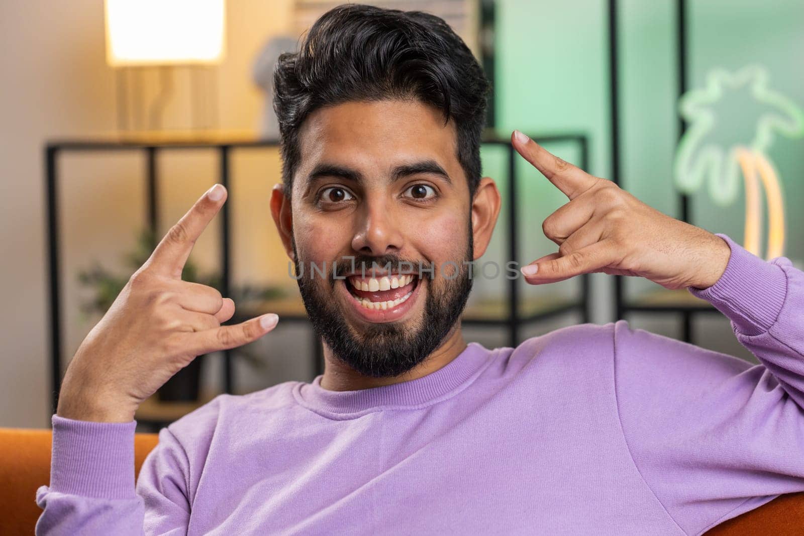 Overjoyed delighted young indian man showing rock n roll gesture by hands, cool sign, shouting yeah with crazy expression, dancing. Hindu guy emotionally rejoicing in success at home living room couch