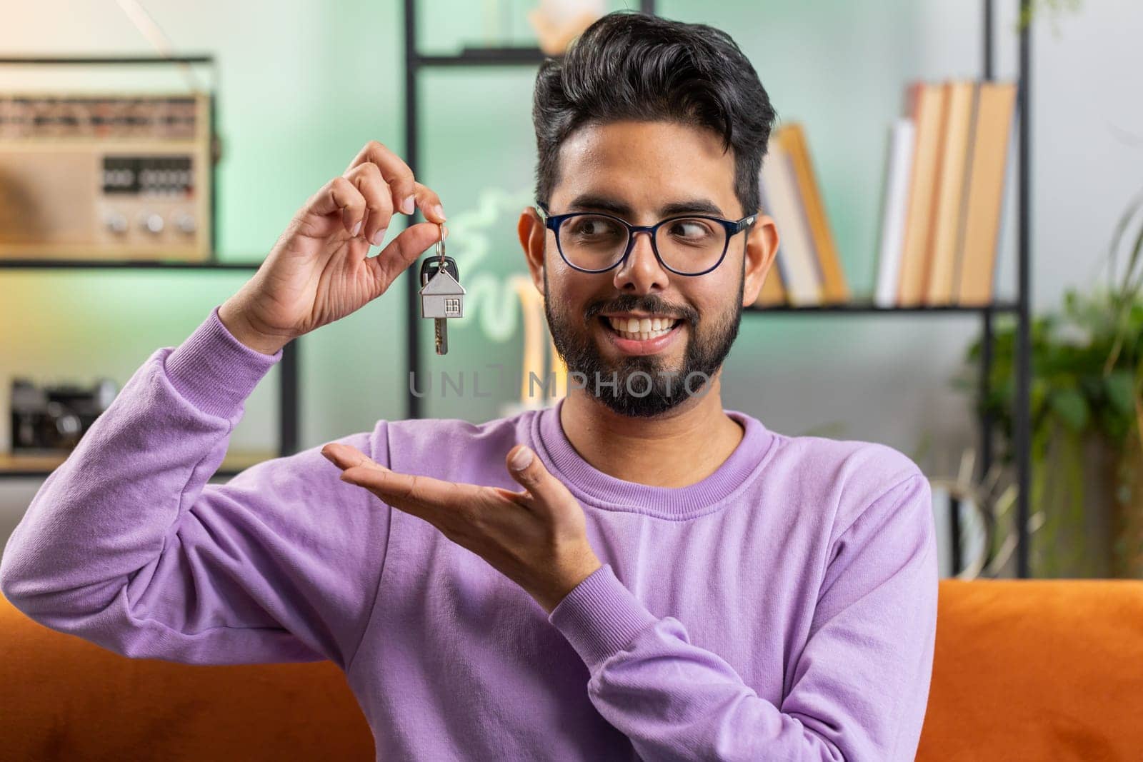 Happy young indian man real estate agent lifting hand showing the keys of new home house apartment, buying or renting property, mortgage loan. Portrait of hindu guy at home apartment room on sofa
