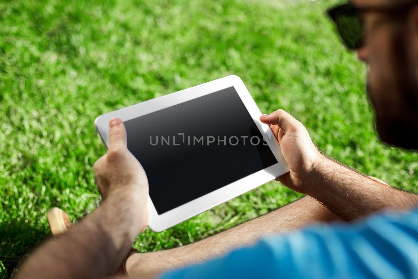 Young man using and typing tablet computer in summer grass. Freelancer working in outdoor park