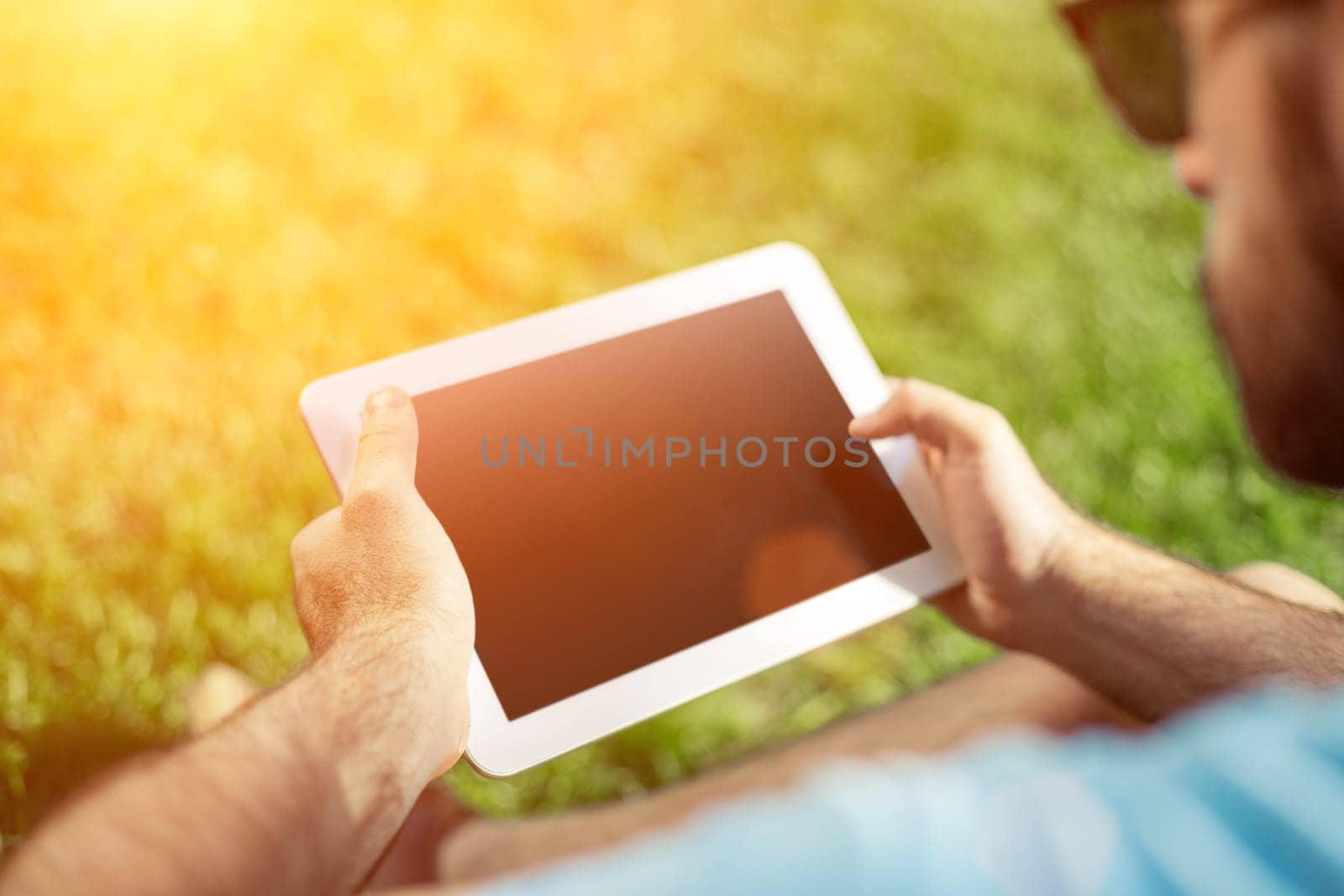 Young man using and typing tablet computer in summer grass. Freelancer working in outdoor park. Sun flare