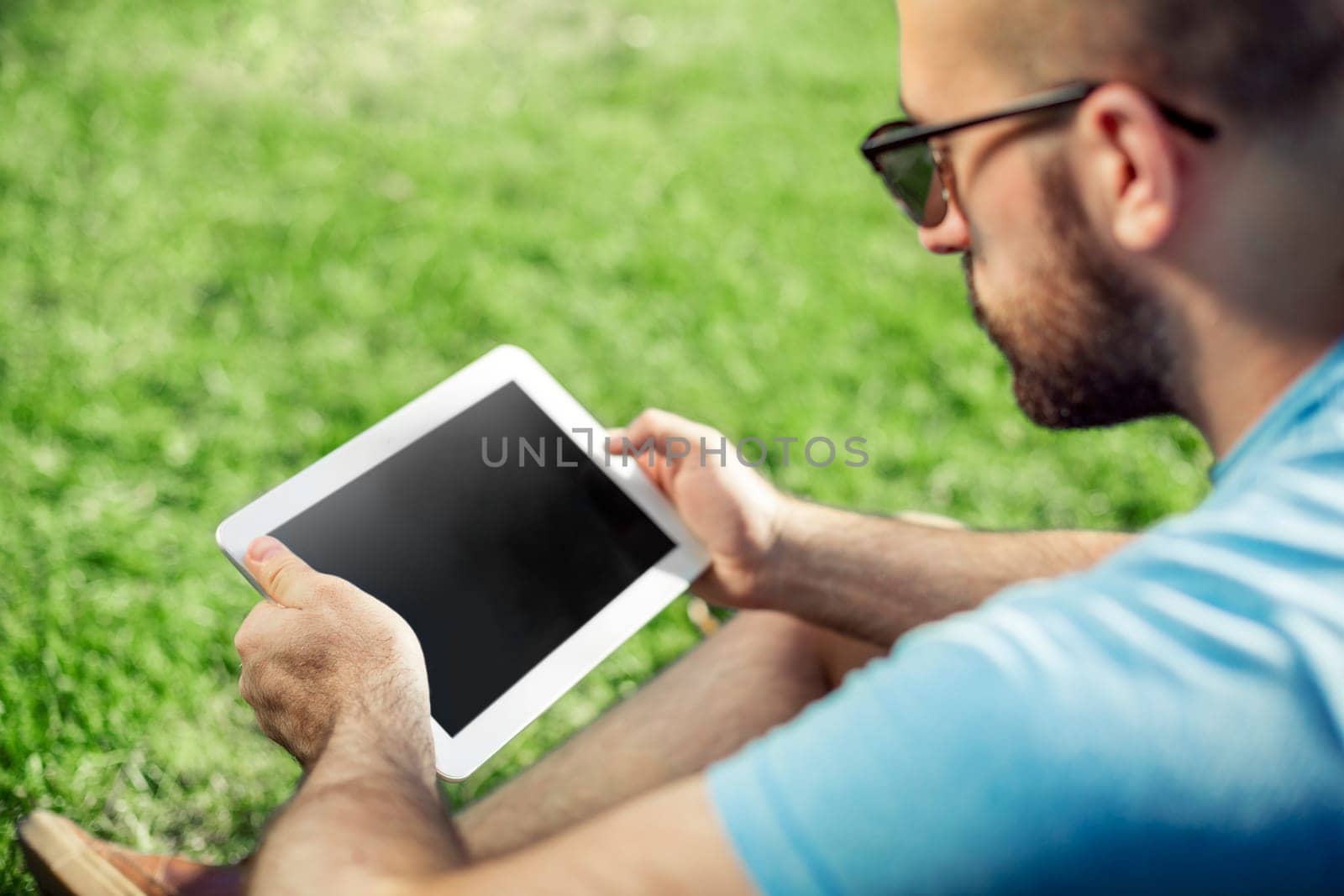Young man using and typing tablet computer in summer grass. Freelancer working in outdoor park