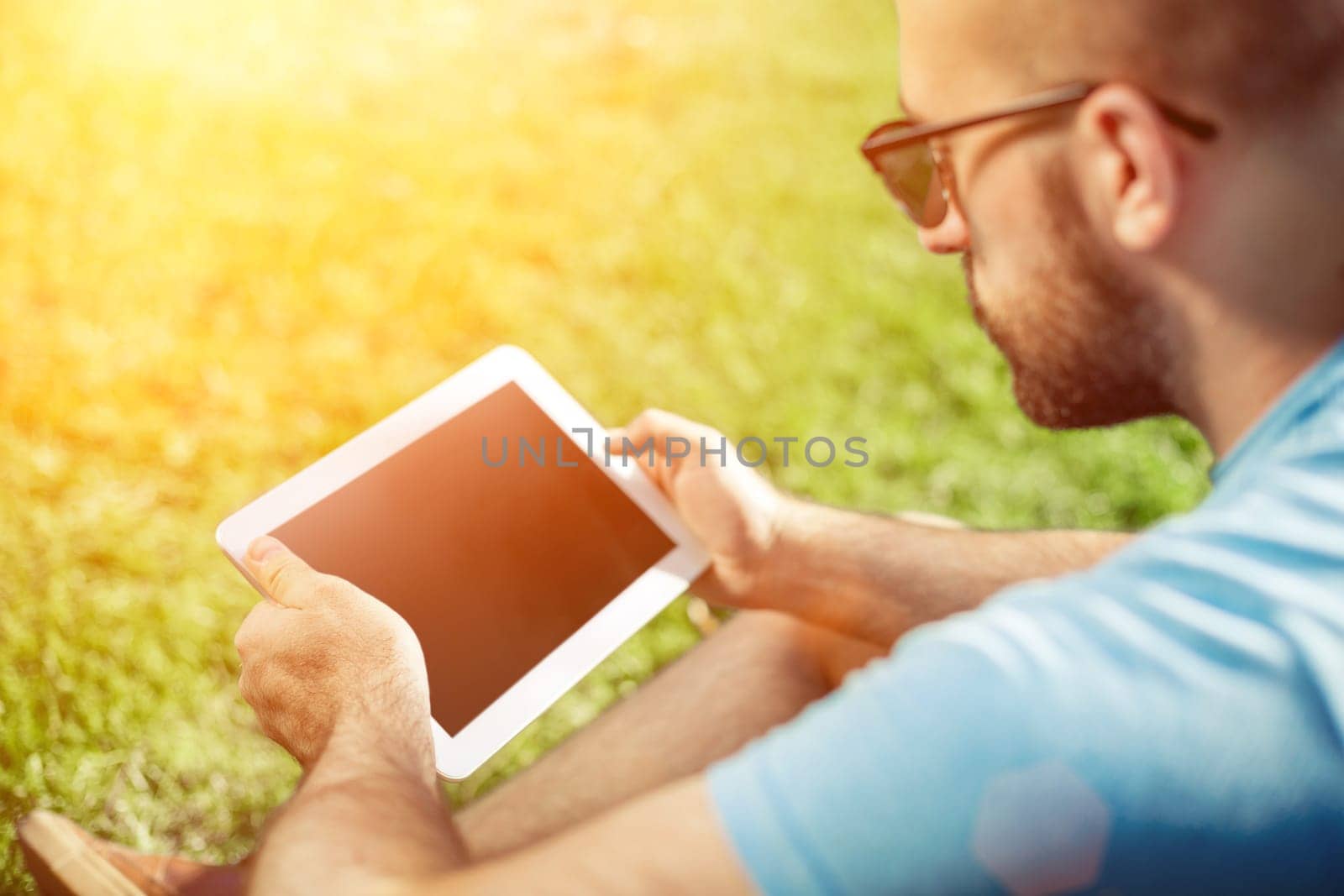 Young man using and typing tablet computer in summer grass. Freelancer working in outdoor park. Sun flare