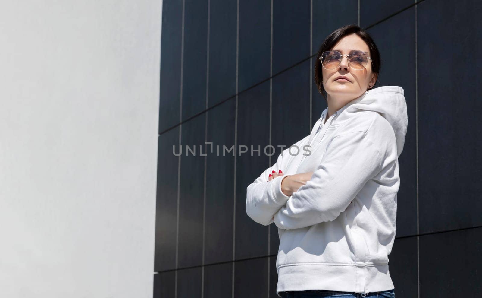 Portrait Mature Brunette Female In White Hoodie and Sunglasses With Crossed Arms, Dark Background Wall. Sportive Confident 40 Yo Beautiful Caucasian Woman Outdoor. Horizontal Plane CopySpace For Text
