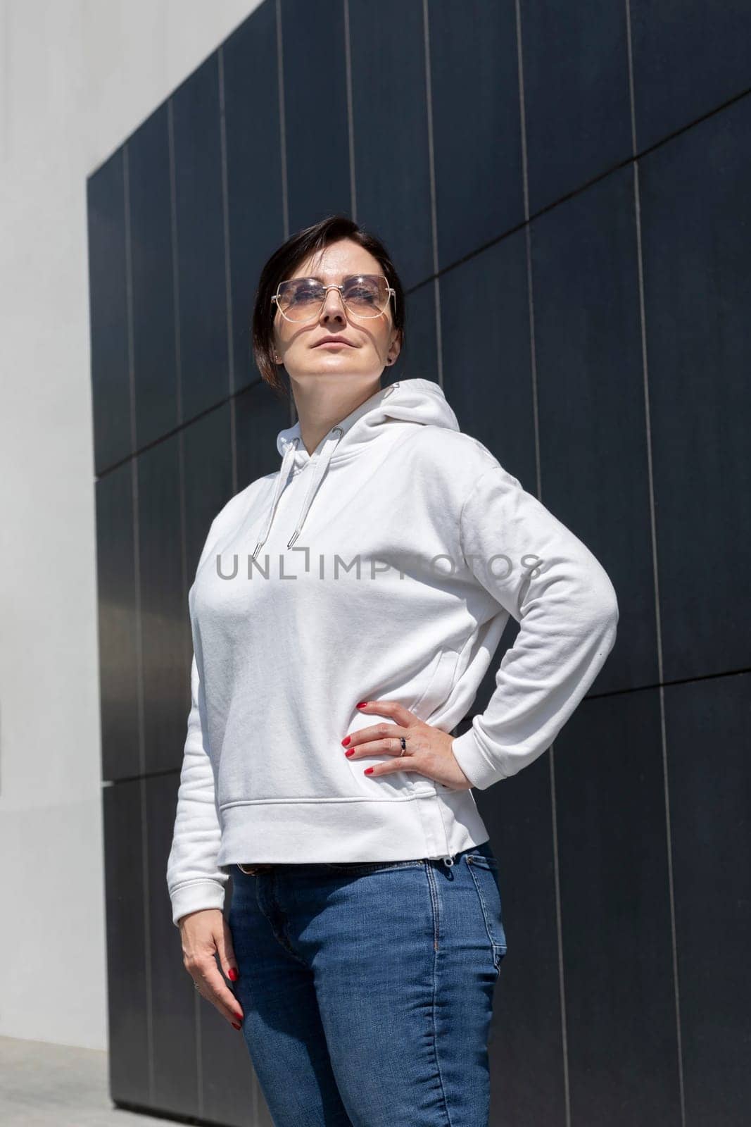 Closeup Pretty Mature Brunette Woman in White Hoody, Jeans and Sunglasses Looks Away On Dark Background Wall. Sportive Confident 40 Yo Beautiful White Woman in Urban Area Vertical Plane. Lifestyle by netatsi