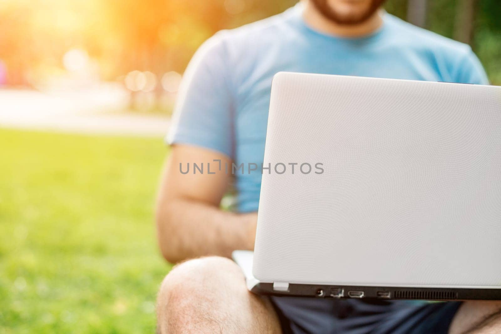 Young man using and typing laptop computer in summer grass. Freelancer working in outdoor park. Sun flare