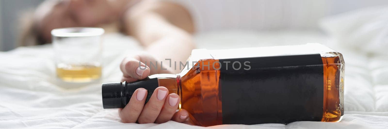 Woman lies in white bed with bottle of whiskey in hand. Alcohol addiction stress and depression concept