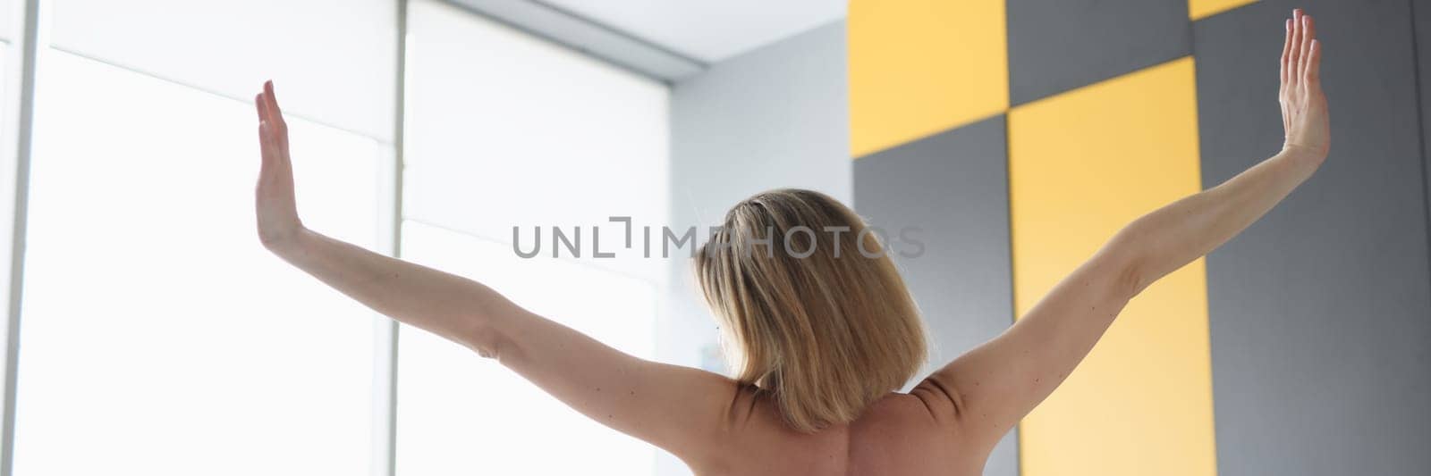 Rear view of naked woman looking out the window and raising her hands up while sitting on bed by kuprevich