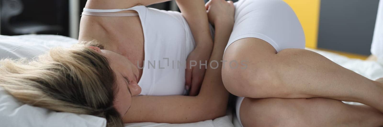Young woman lies in bed and holds on to sore stomach in bedroom by kuprevich