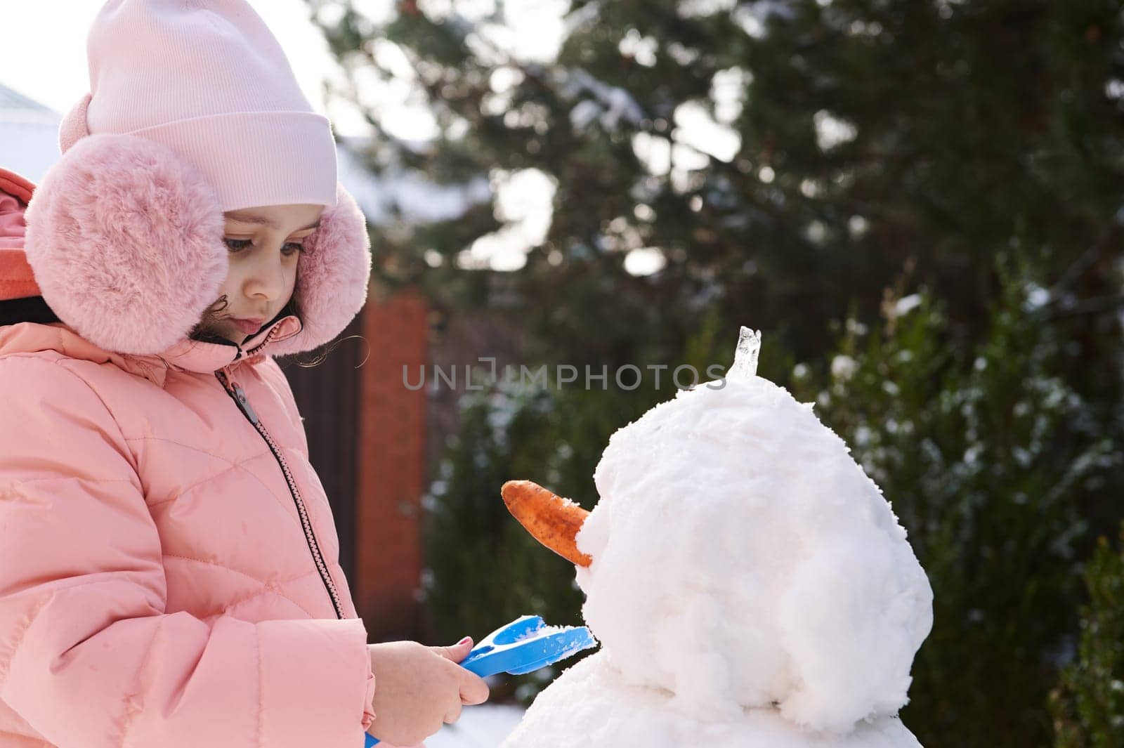 Close-up portrait lovely little child girl in pink down jacket and fluffy earmuffs, building snowman in snowy backyard by artgf
