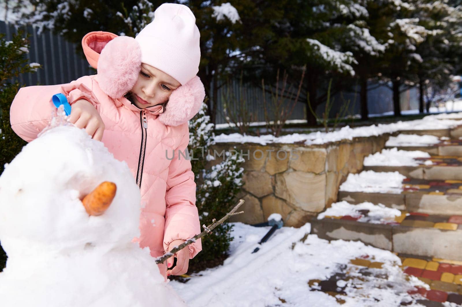 Caucasian lovely little kid girl 5 years old, dressed in pink down jacket and stylish fluffy earmuffs holding a shovel and building a snowman outdoors. Leisure activities and games at wintertime