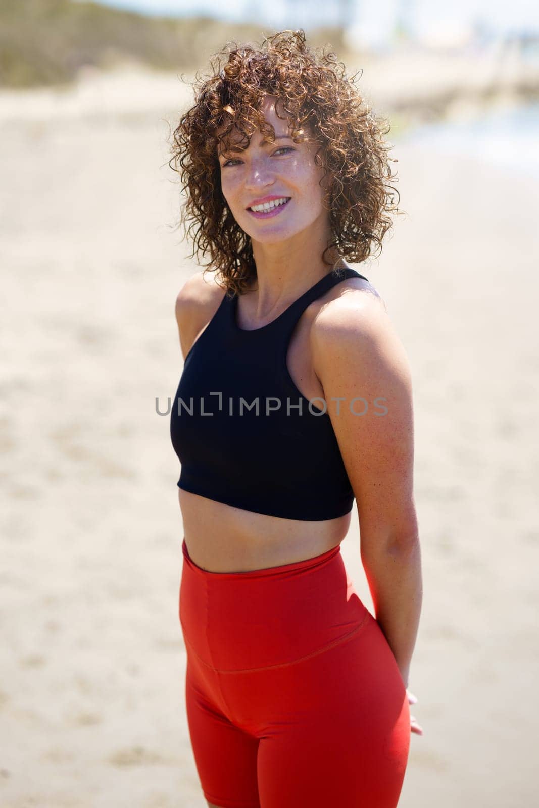Positive young female athlete with curly hair in top and leggings standing on sandy beach with hands behind back and looking at camera