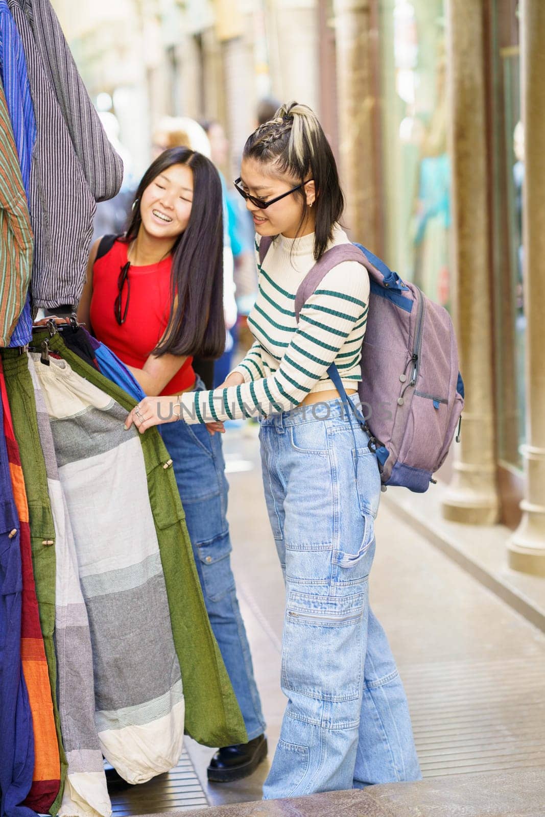 Positive young Asian women, in casual clothes with backpack smiling and looking at camera while shopping clothes on street market in city during vacation