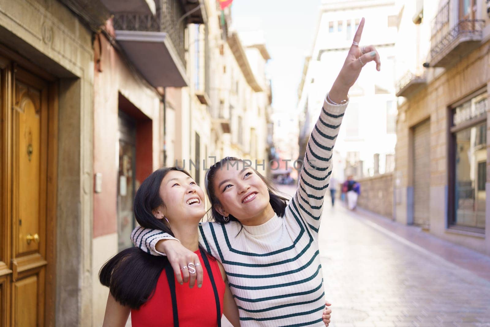 Happy Asian girlfriends in casual clothes walking on street of old town and viewing buildings while pointing up and hugging on sunny day