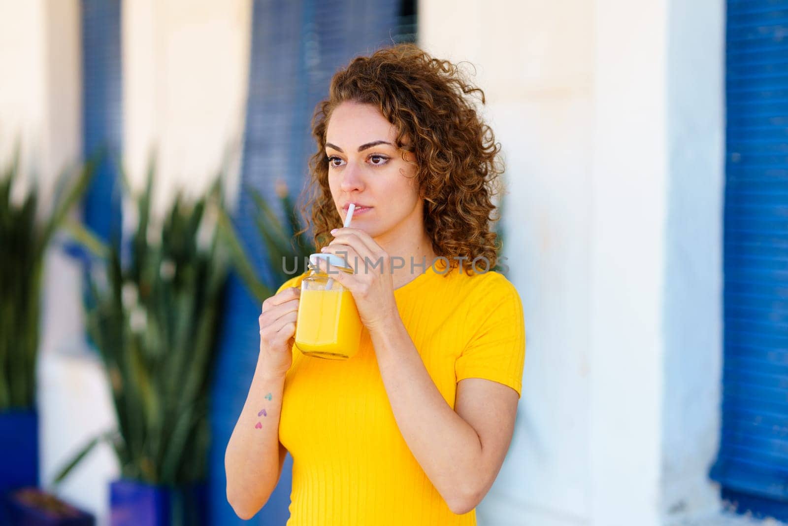 Pensive young female in yellow t shirt with curly brown hair looking away and drinking fresh juice from glass jar with straw on blurred street