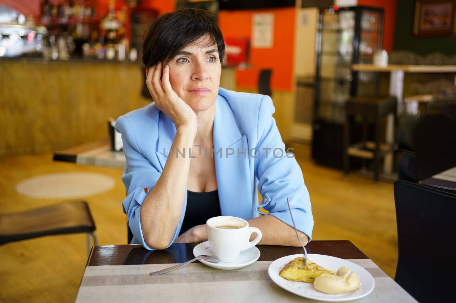 Thoughtful lady in elegant jacket sitting at table with cup of beverage and piece of cake while leaning on arm in cafeteria