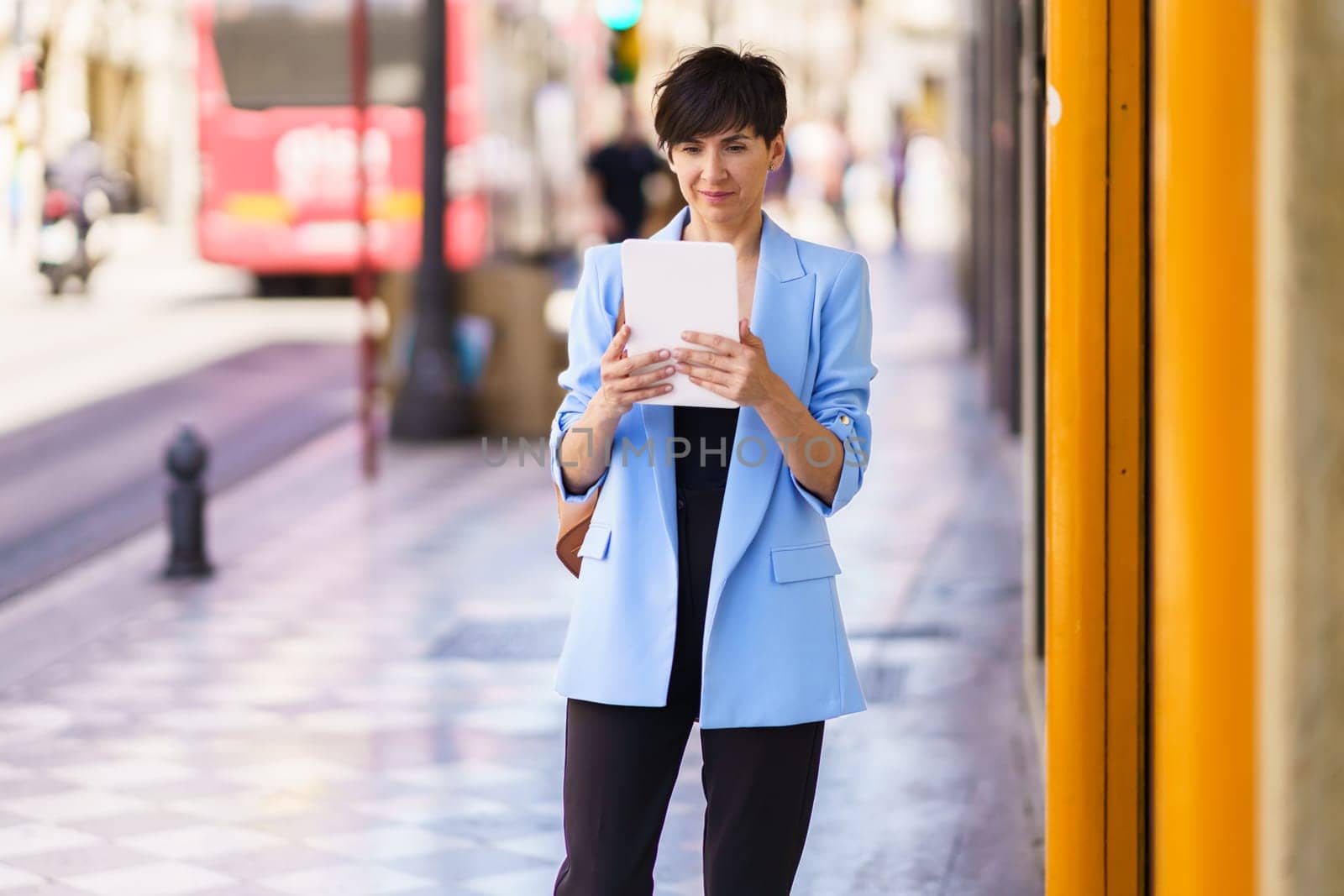 Concentrated female entrepreneur in formal clothes, reading on tablet while standing on city street and looking at screen against blurred background of people and bus in daylight