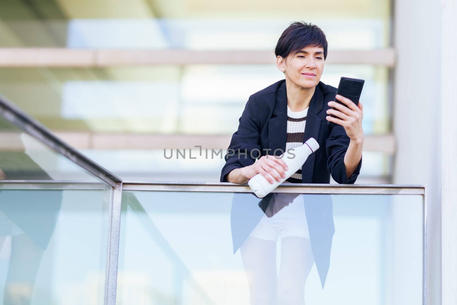 Smiling female in black jacket holding hand bottle while surfing internet on mobile phone and leaning against glass wall of building with transparent wall