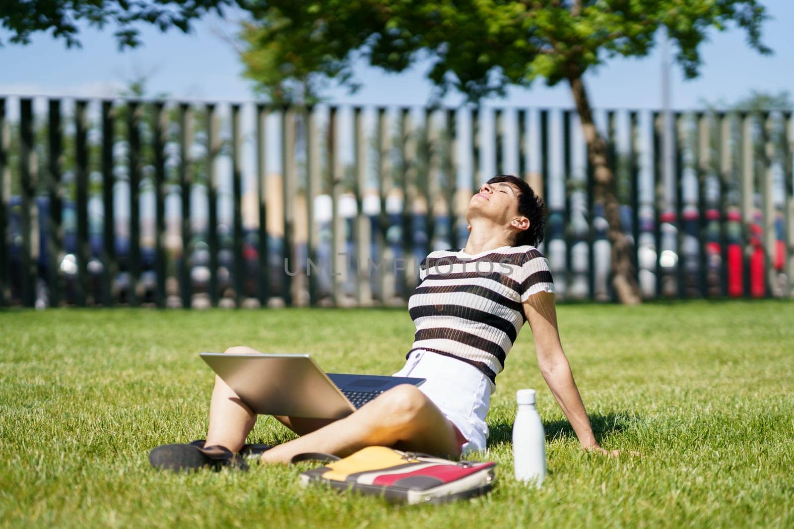 Female blogger sitting on green lawn with laptop and enjoying fresh air while relaxing after using online studies against blurred background