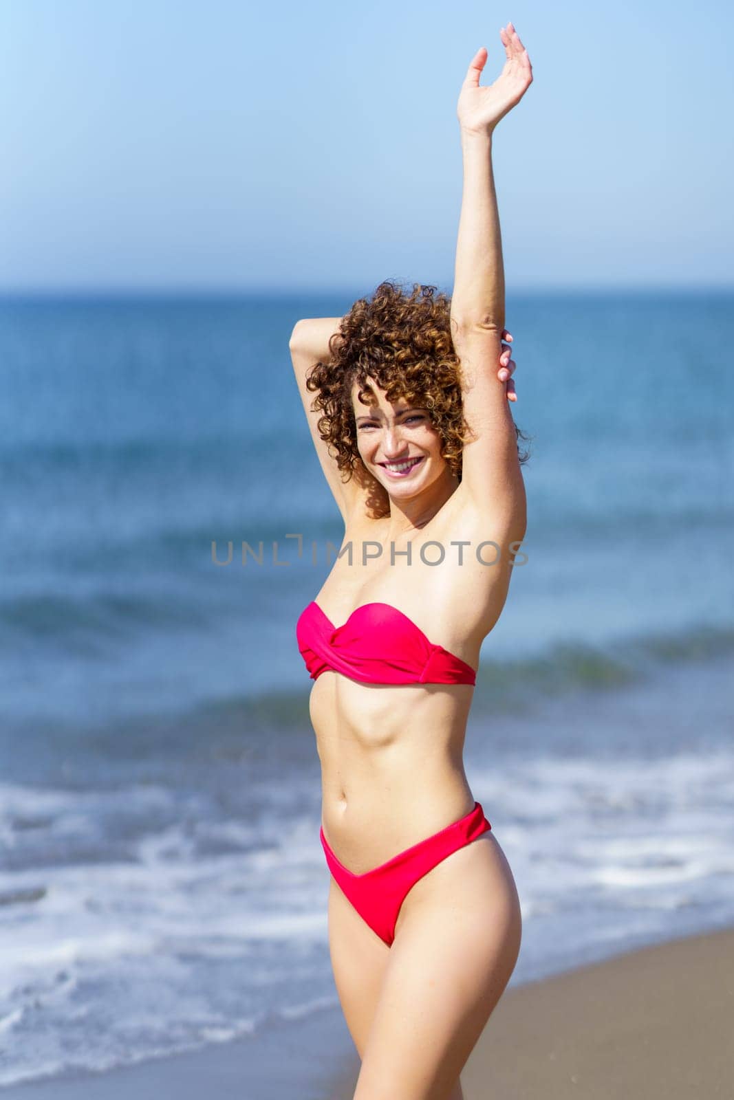 Cheerful young female redhead in pink bikini standing and touching outstretched arm with hand from behind head, while looking at camera in sunlight against blue seawater and sky