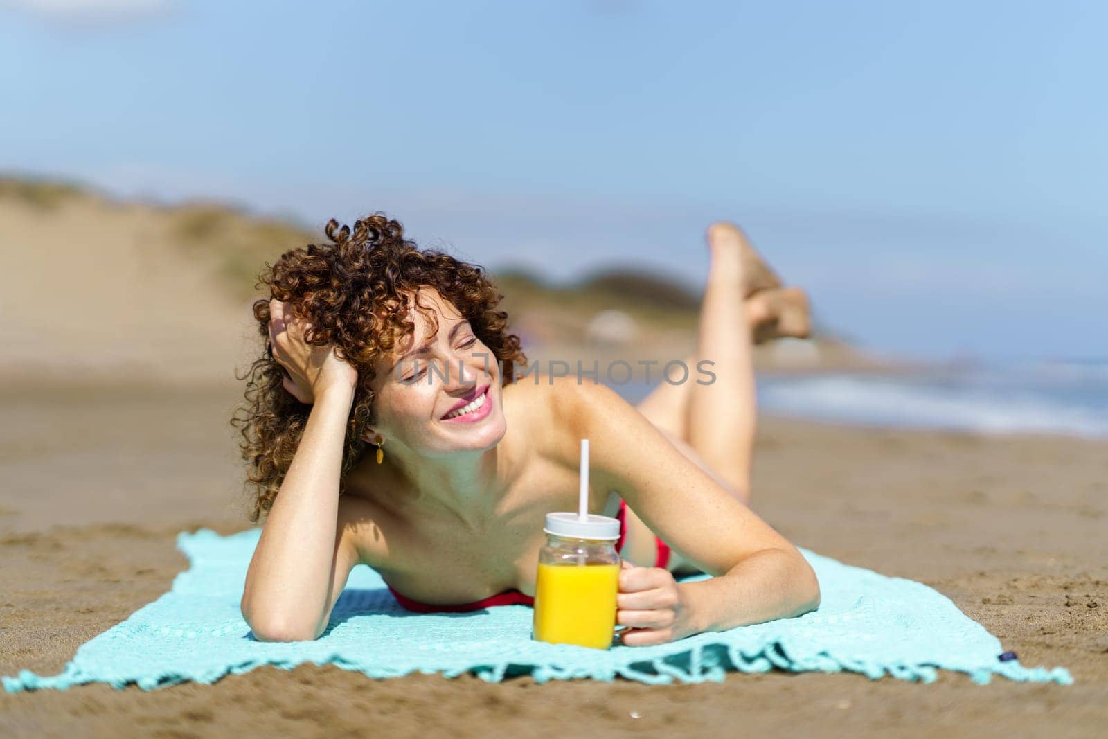 Cheerful young woman with curly hair smiling and closing eyes in sunlight while suntanning and drinking juice on sandy beach