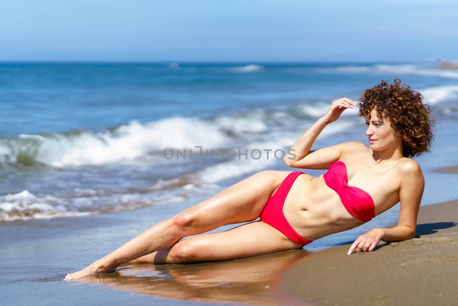 Dreamy young woman with curly hair and in bikini lying on sandy shore of ocean in sunlight looking away