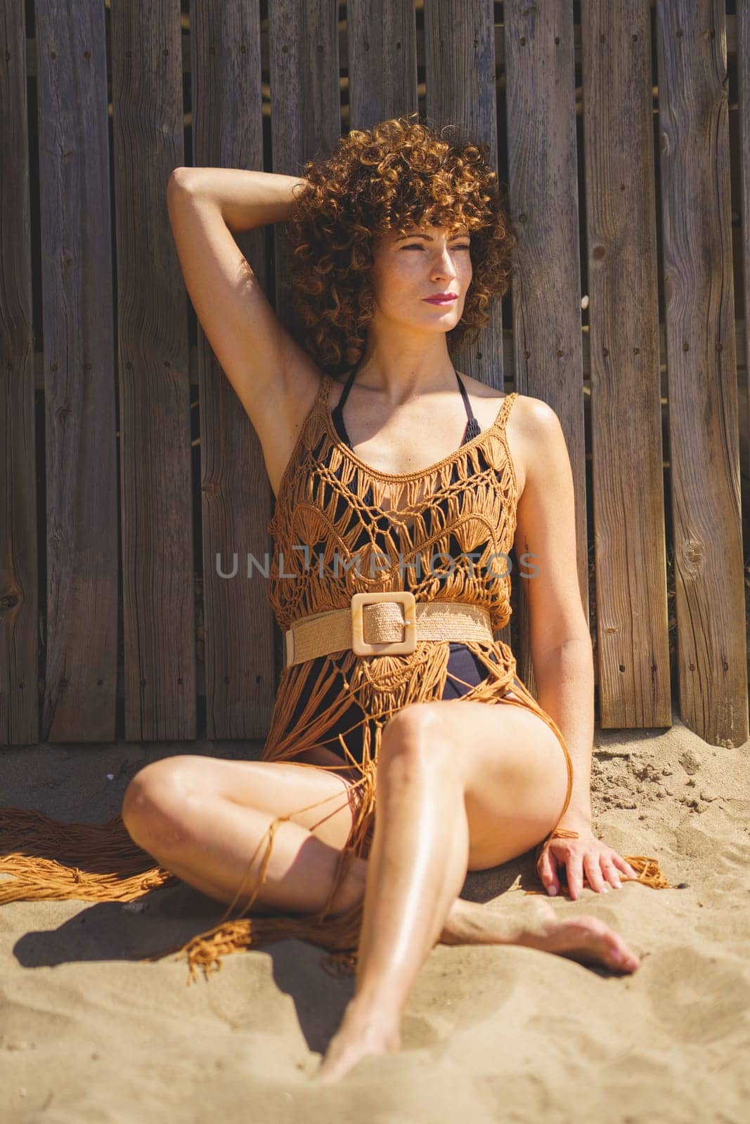 Full body of young barefoot female wearing swimwear and brown knitted beach cape sitting on sandy coast, with hand behind head in sunlight while looking away thoughtfully