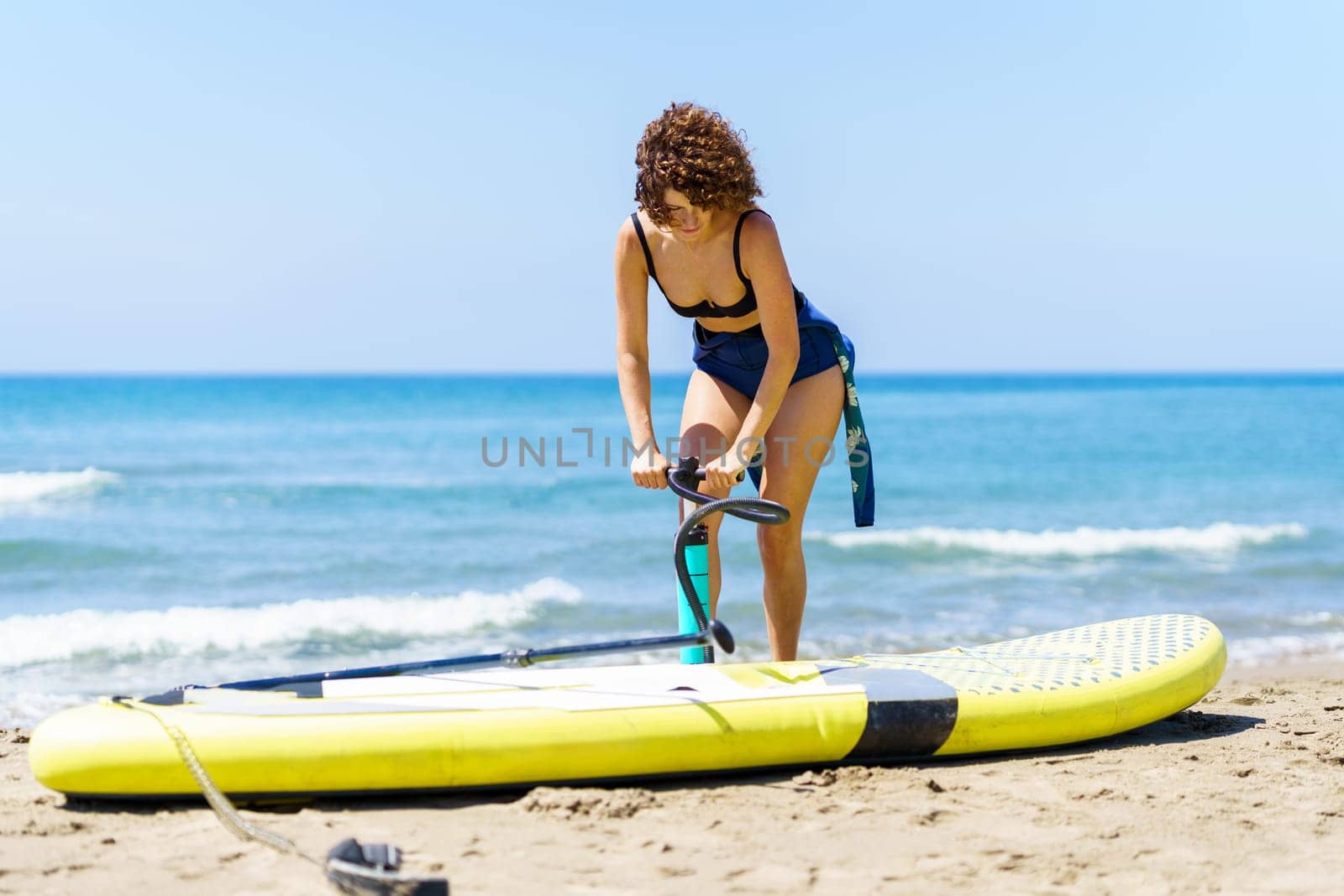 Focused young female with curly red hair in swimwear standing near paddleboard and pumping air, with handpump while looking down against blurred waving sea and blue sky